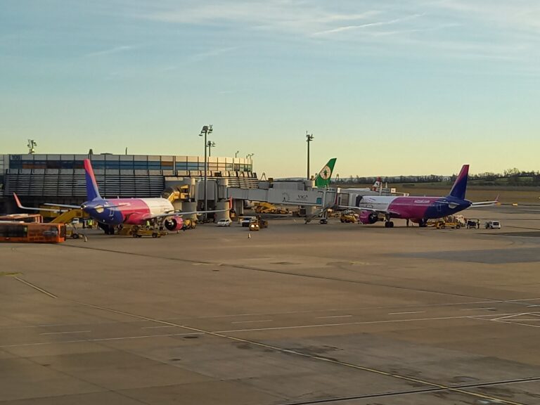 Dvě letadla Wizz Air v růžovo-fialových barvách stojí u nástupních mostů, v pozadí budova terminálu
