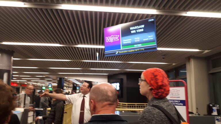 Pracovník v košili a kravatě ukazuje cestujícím směr v odletové hale u obrazovky s logem Wizz Air